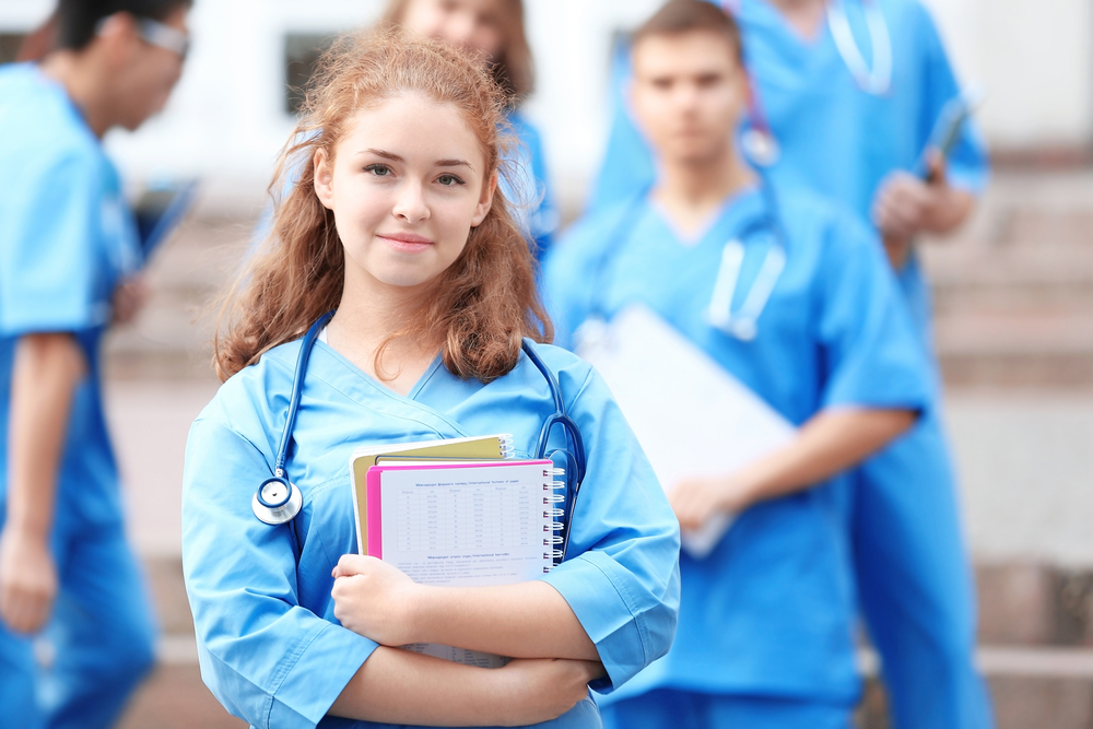 Young female nursing student in scrubs with a stethoscope holding notebooks and smiling at the camera.